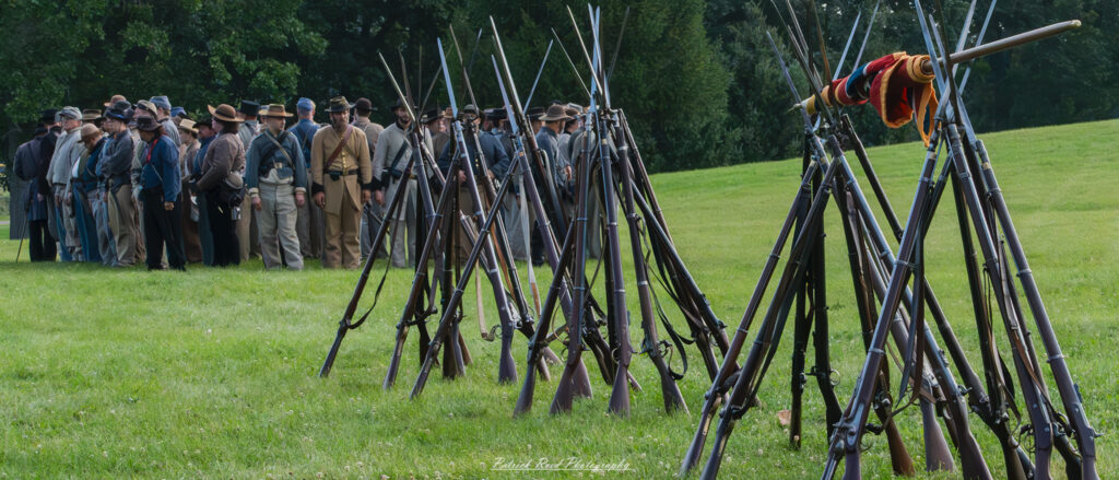A group of Confederate soldiers in a field, gathered around with their weapons nearby. The scene shows the soldiers in uniform, perhaps taking a brief rest or preparing for action. The surrounding field and scattered equipment highlight the rugged and often makeshift nature of wartime life.